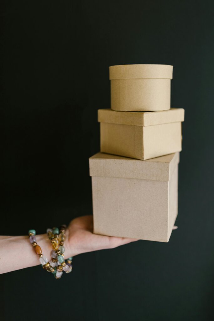 pexels-photo-7309438-7309438 Close-up of kraft boxes held by a hand with beaded bracelets on a dark background.